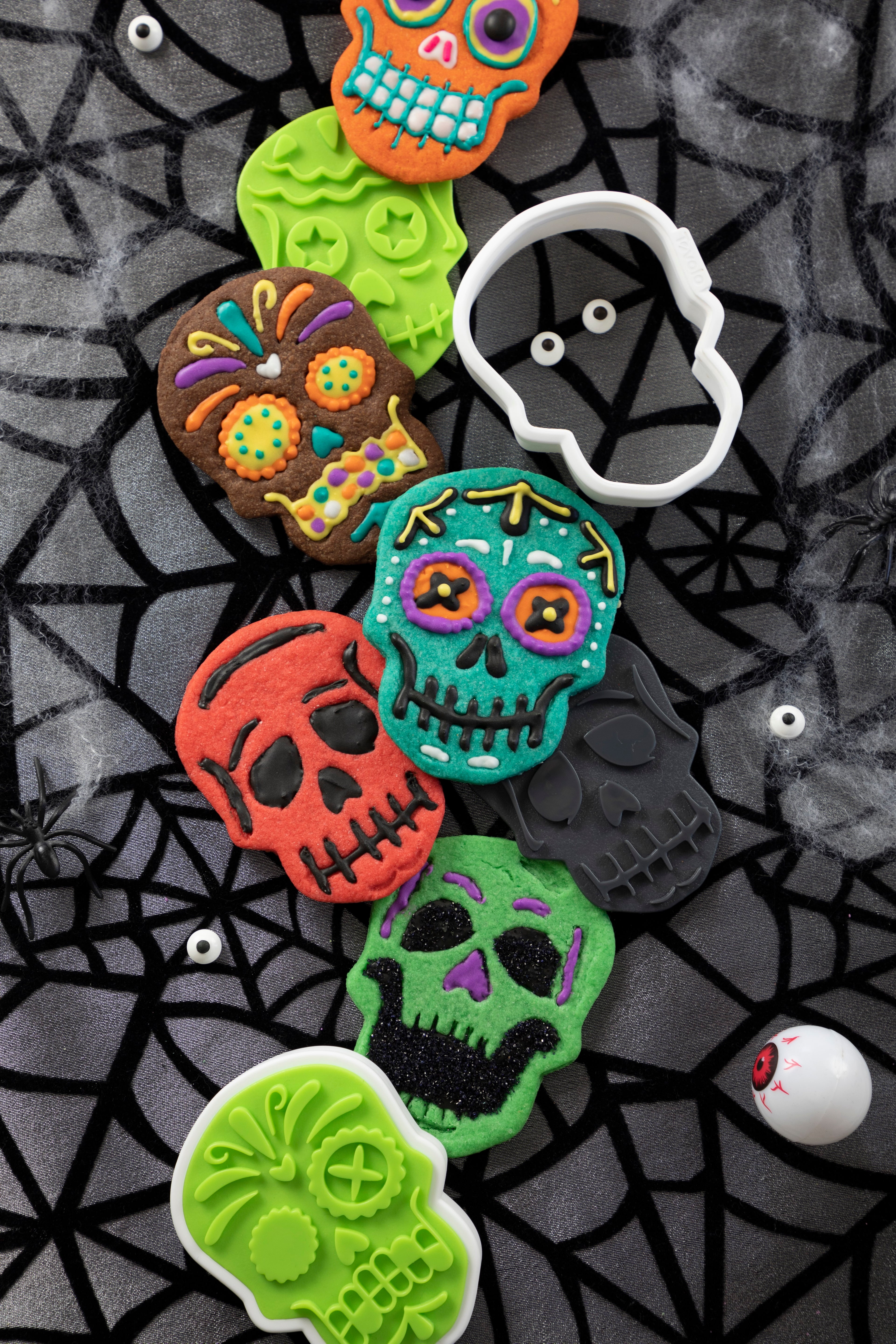 Colorful skull cookies made with Skull Cookie Cutters are decorated with icing and sprinkles on a spiderweb-patterned surface, surrounded by fake eyeballs, spiders, and a white skull-shaped cookie cutter.