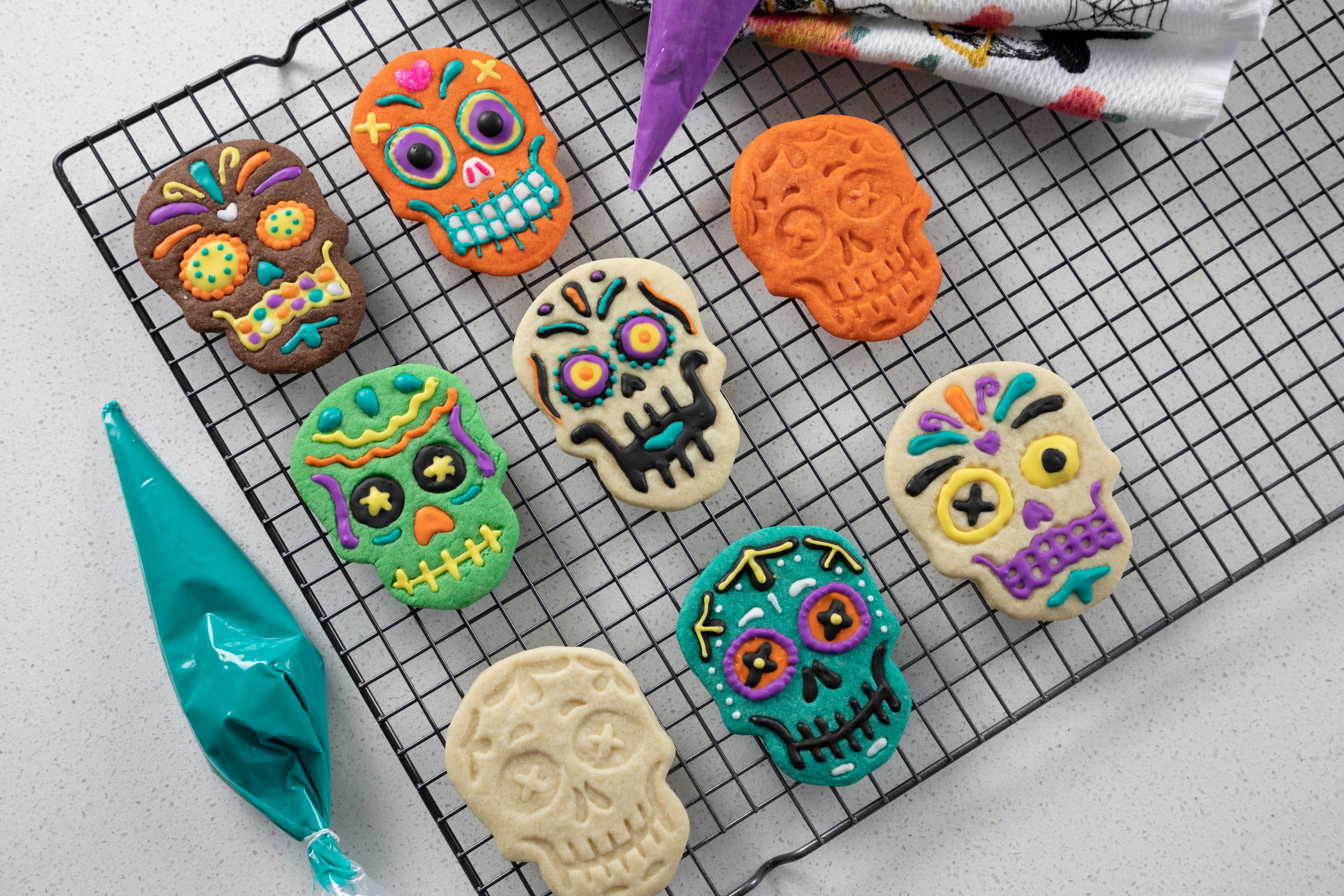 Sugar skull-shaped cookies, made with Skull Cookie Cutters, rest on a cooling rack, their bright icing reminiscent of festive Christmas cookies. A nearby piping bag of blue icing is ready to add more creative designs.