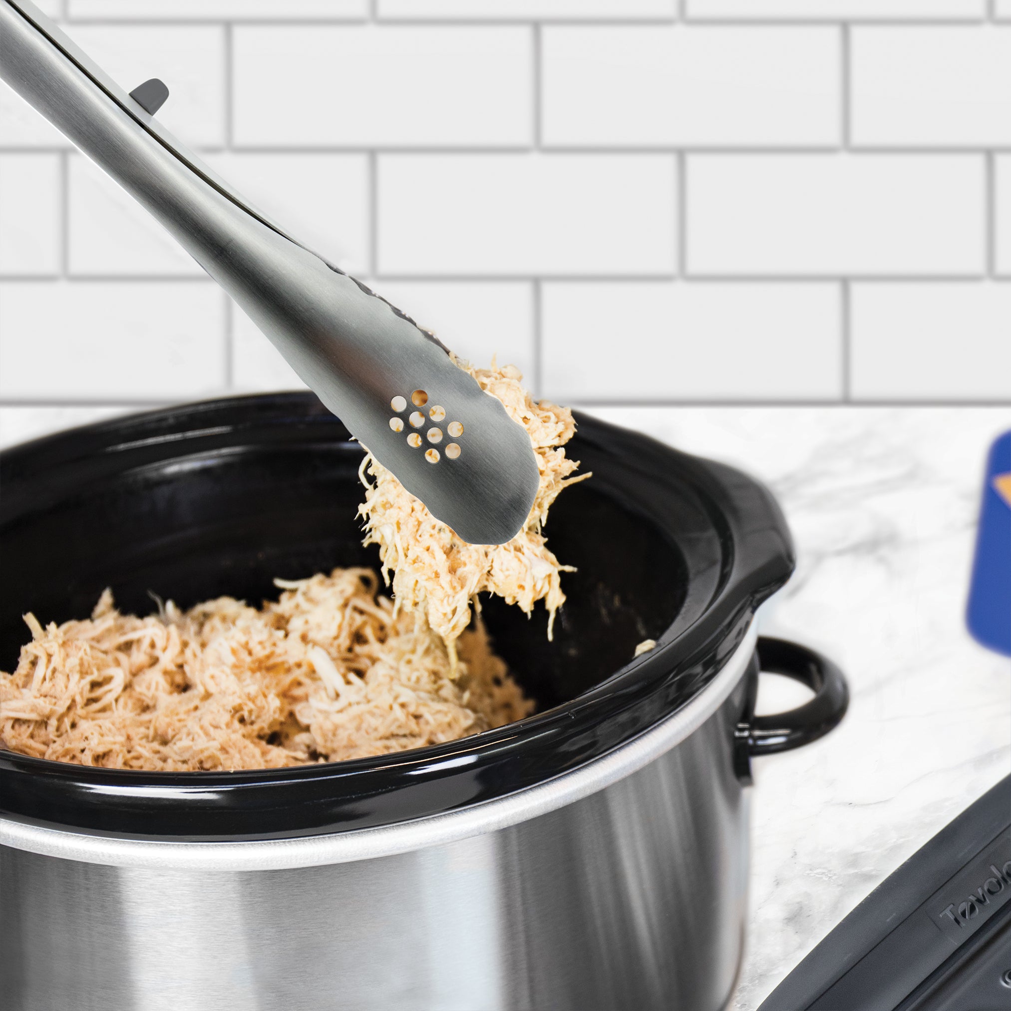 A pair of Stainless Steel Tongs holds shredded chicken above a black and silver slow cooker filled with more chicken, making the Stainless Steel Tongs perfect for meal prep. A white tiled wall is visible in the background.