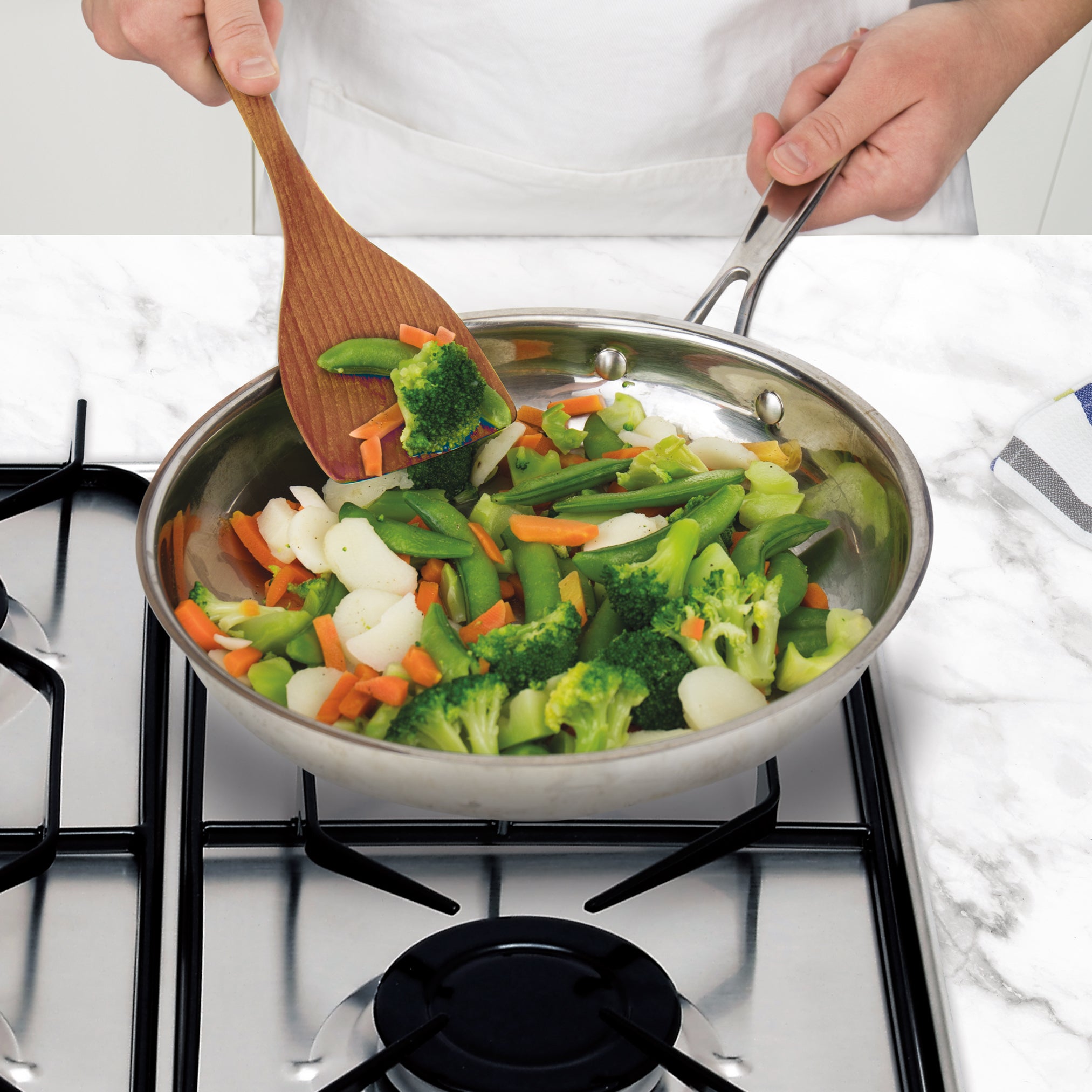 A person cooking mixed vegetables—broccoli, carrots, snap peas, and water chestnuts—in a stainless steel pan on the stove, stirring with Olivewood Utensils.