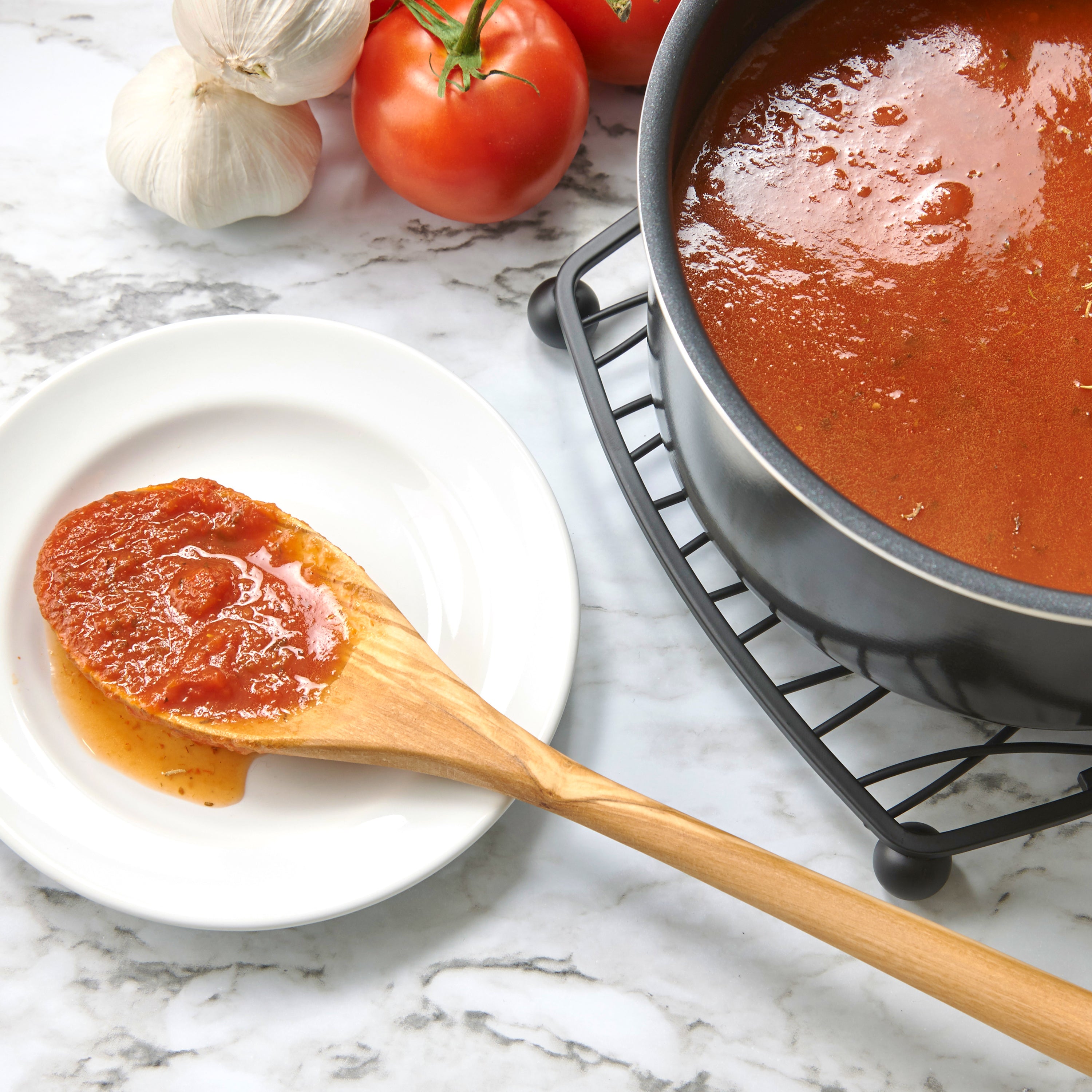 An Olivewood Utensil coated in tomato sauce rests on a white plate beside a pot of sauce, with fresh tomatoes and garlic arranged on a marble countertop.