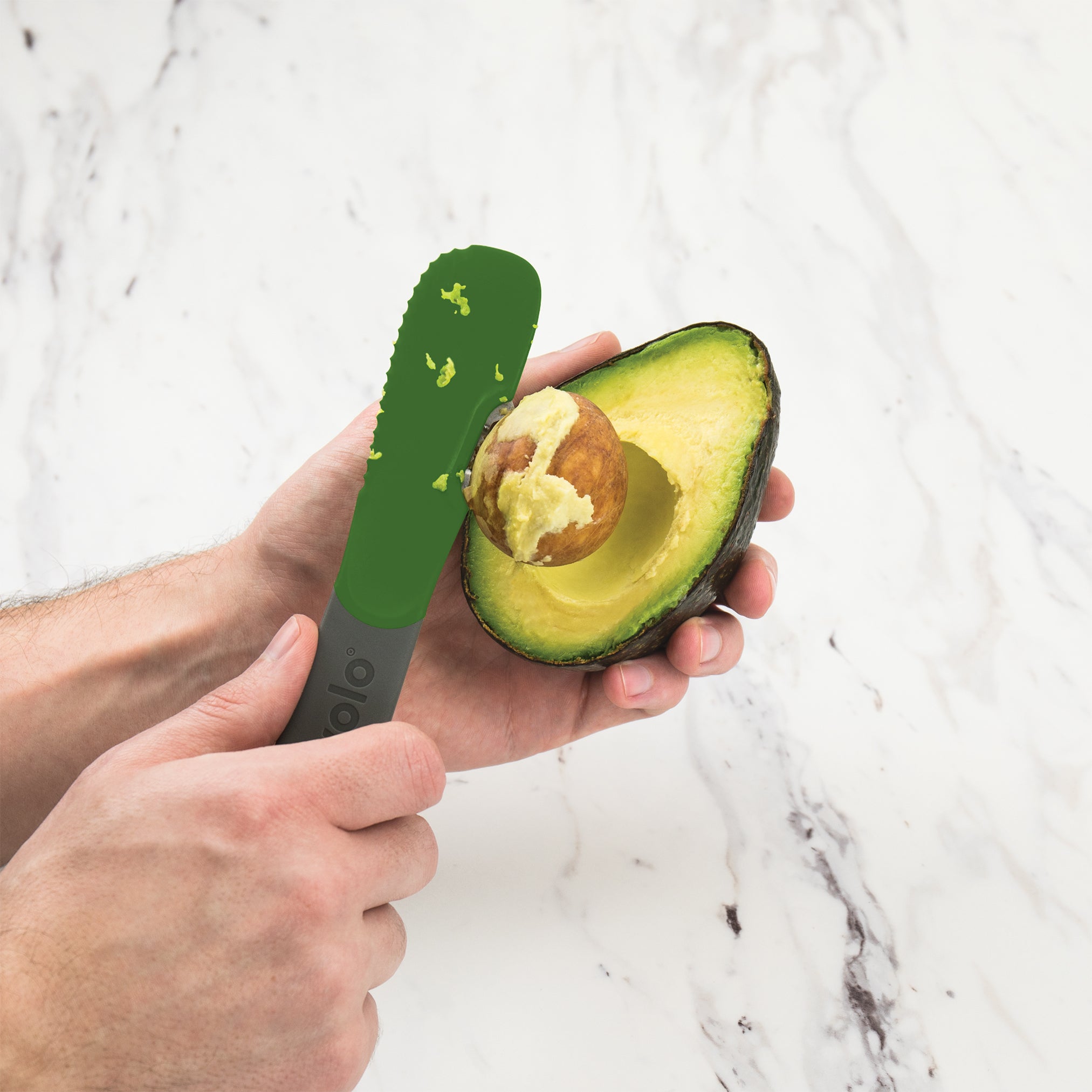 Using the 3-in-1 Avocado Tool, someone removes the pit from a halved avocado on a white marble surface.