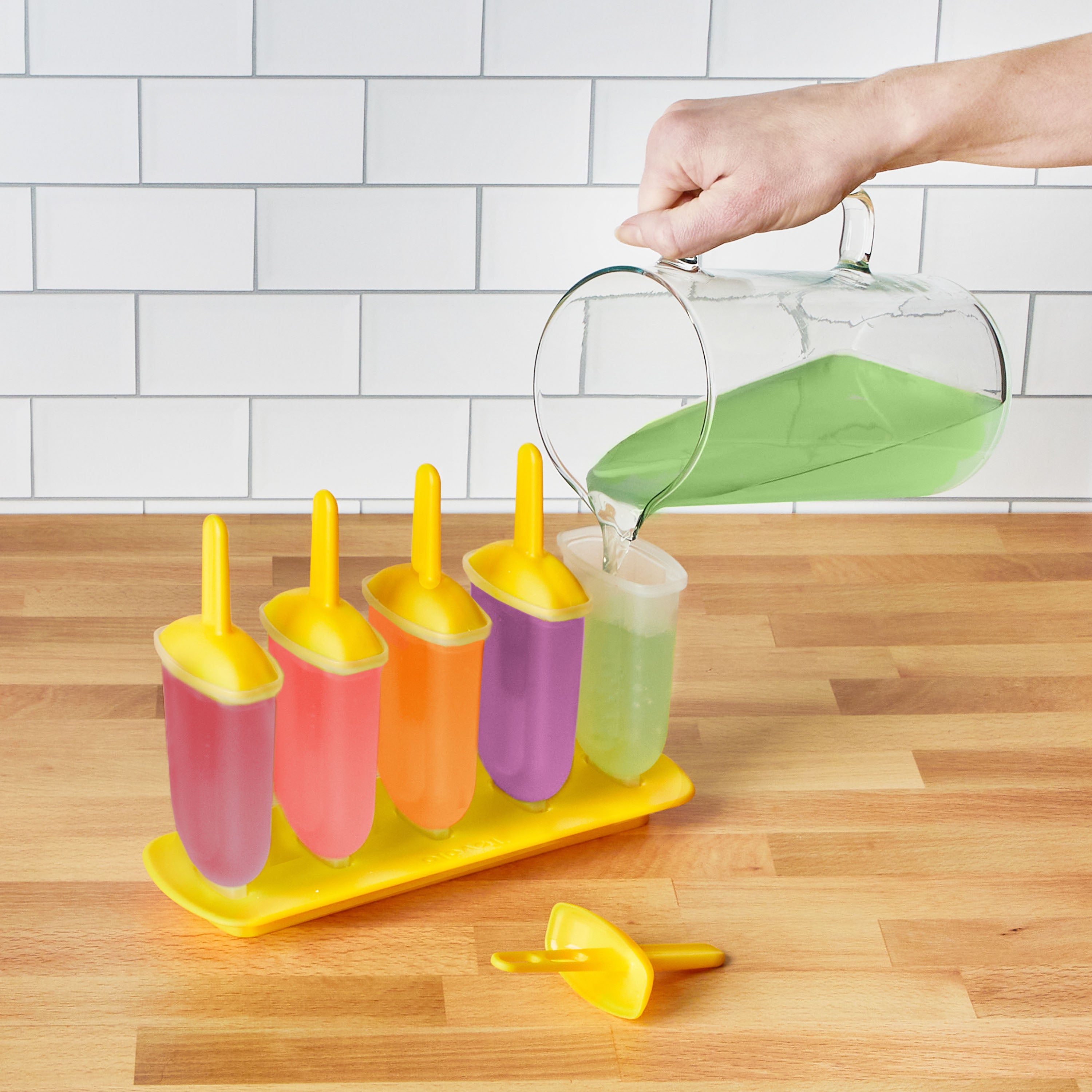 A hand pours green liquid from a pitcher into Pop Molds next to three filled with pink, orange, and purple on a slimline tray atop a wooden counter—perfect for making homemade frozen treats.