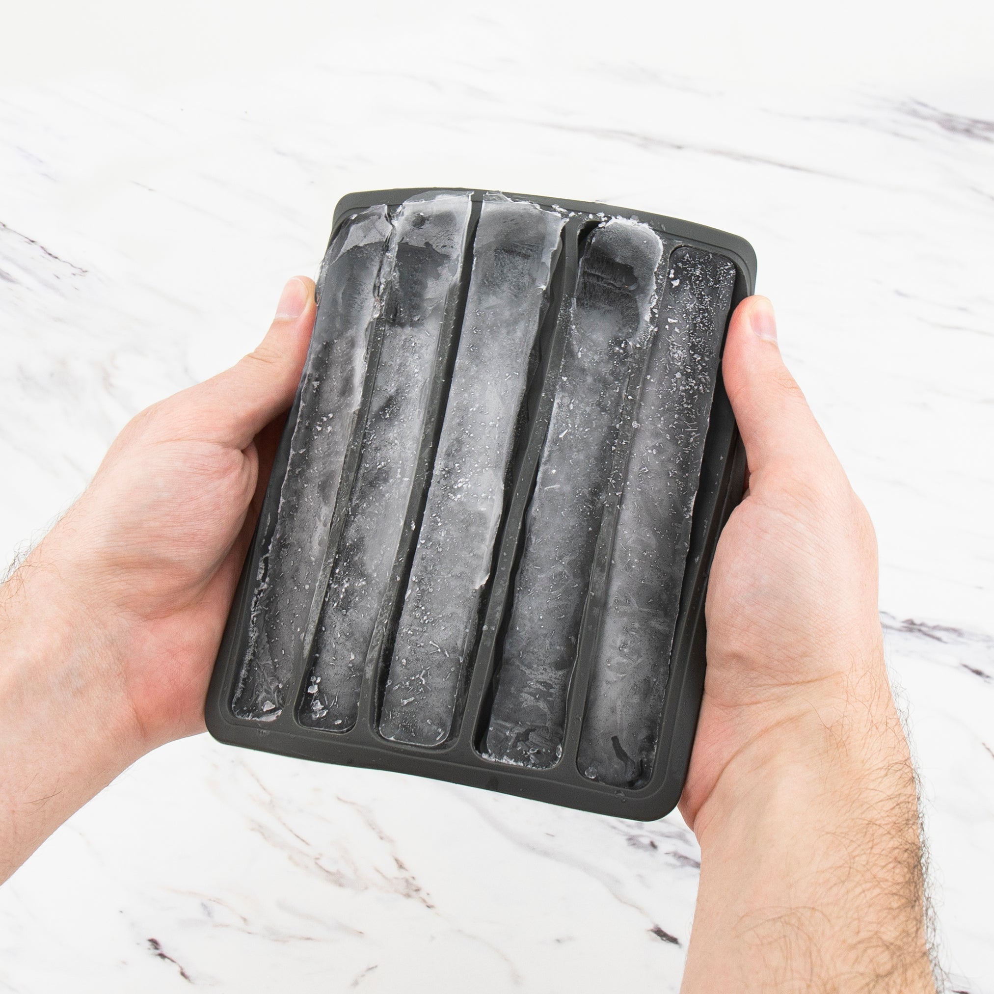 Someone holds a Water Bottle Ice Mold, a black silicone tray that makes four long, narrow ice sticks, over a white marble surface.