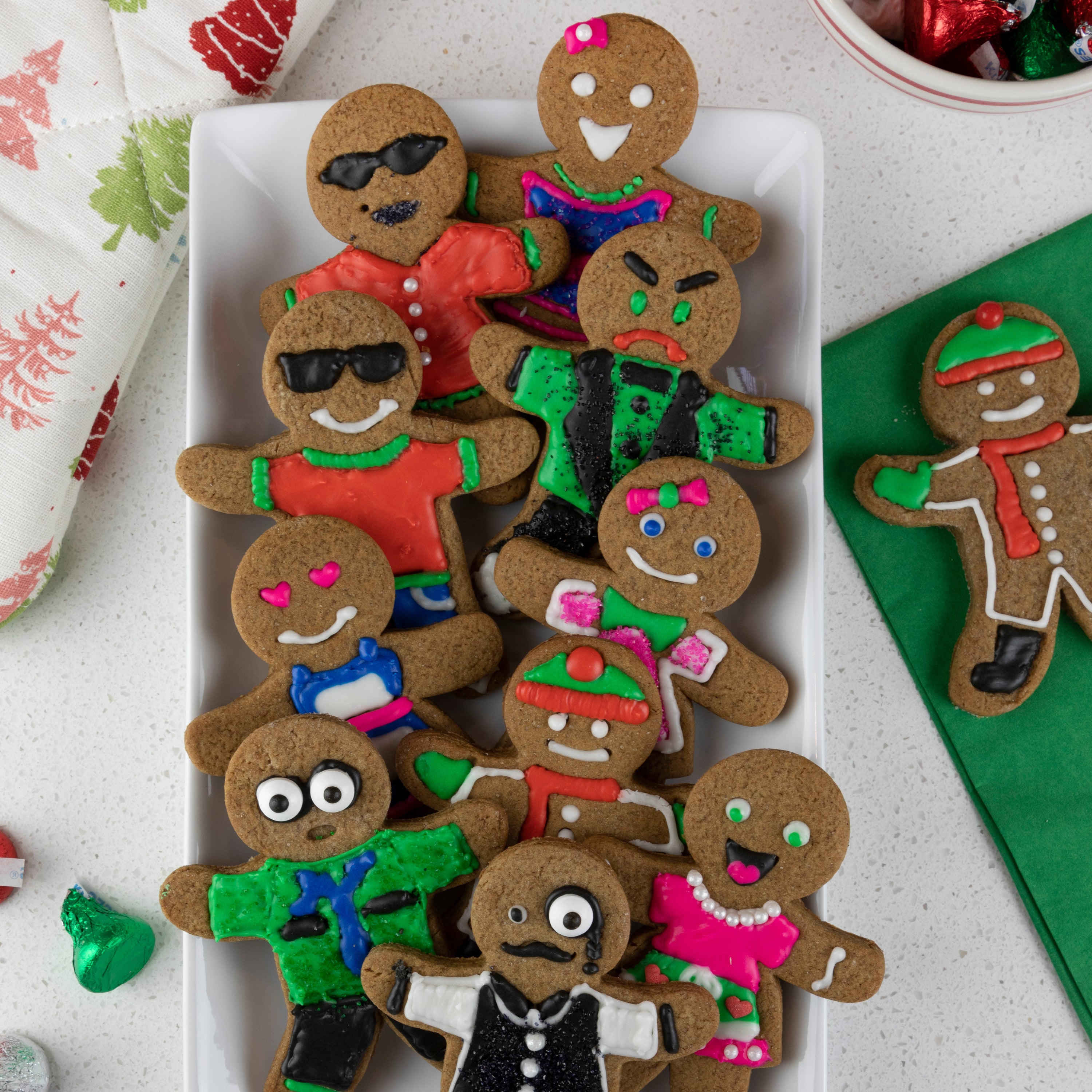 A rectangular plate displays Christmas cookies shaped like people, each with colorful icing outfits and expressions, made using Gingerbread Cookie Cutters. A green napkin and festive baking items surround the plate.