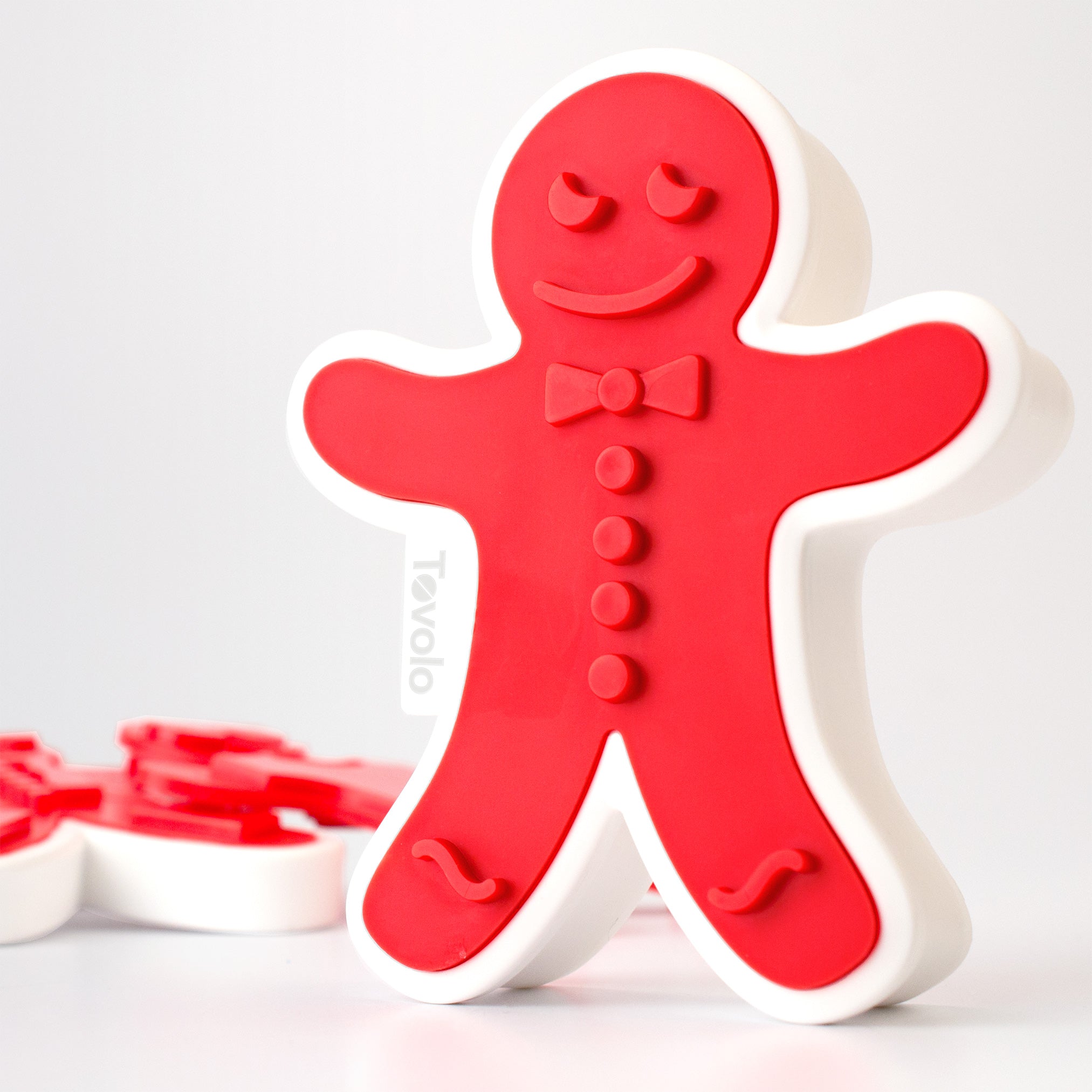 The Gingerbread Cookie Cutters, shaped like gingerbread men with red and white details, smiles, bow ties, and buttons, stand upright against a white background—ideal for winter baking. Similar Christmas cookie cutters appear blurred in the background.