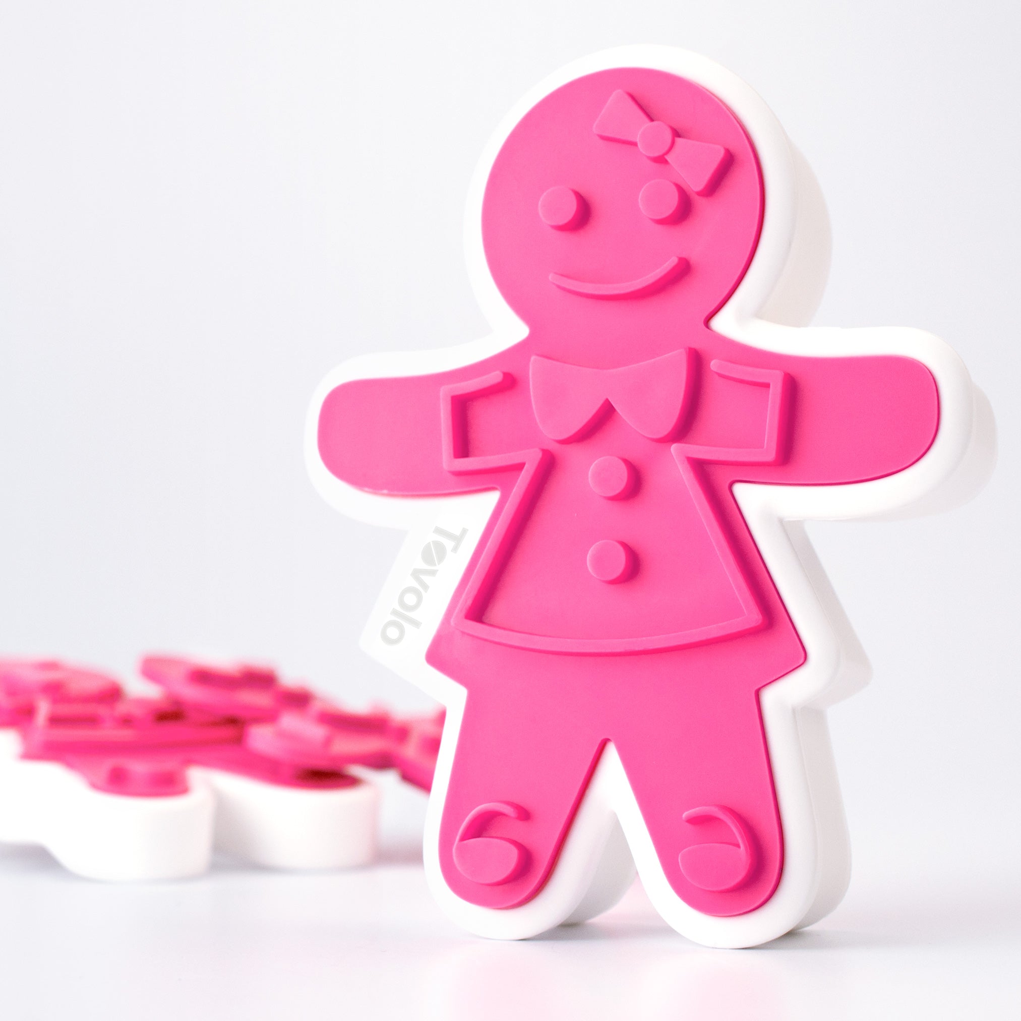 A pink and white Gingerbread Cookie Cutters girl with a bow and buttons stands upright on a white surface, perfect for making festive Christmas cookies, with additional cookie cutters blurred in the background.