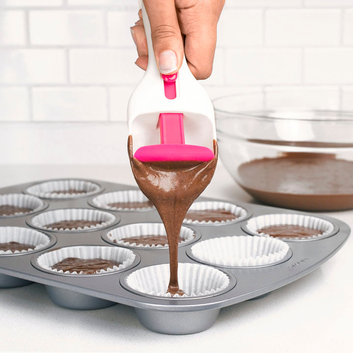 A hand uses the Cupcake Scoop to pour chocolate cake batter into paper-lined muffin cups, while a glass bowl with extra batter sits on the kitchen counter in the background.