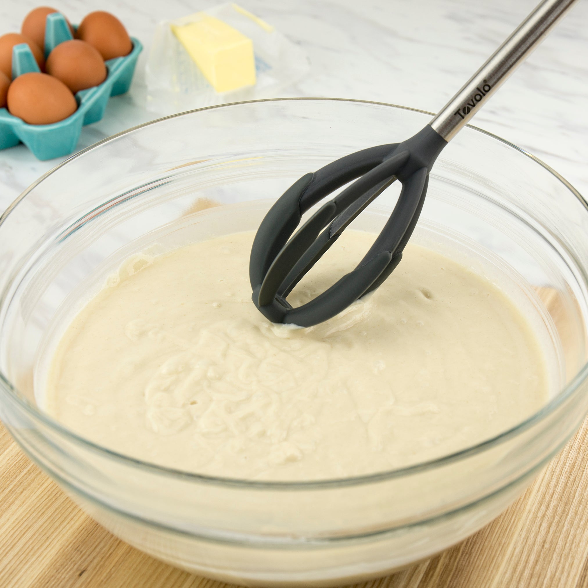 A glass bowl with cake batter is being mixed using the Better Batter Tool. In the background, a carton of brown eggs and a stick of butter sit on the countertop.