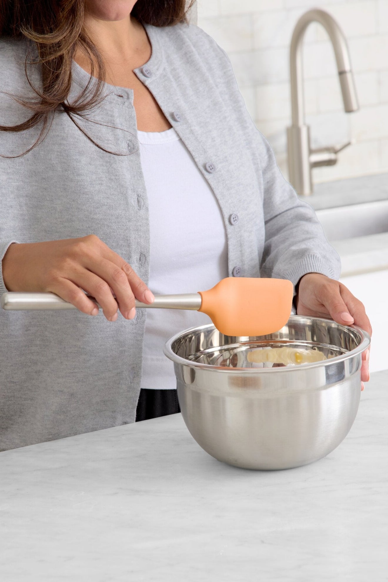 A woman in a gray cardigan uses an orange spatula to mix ingredients in a stainless steel bowl on a kitchen counter.