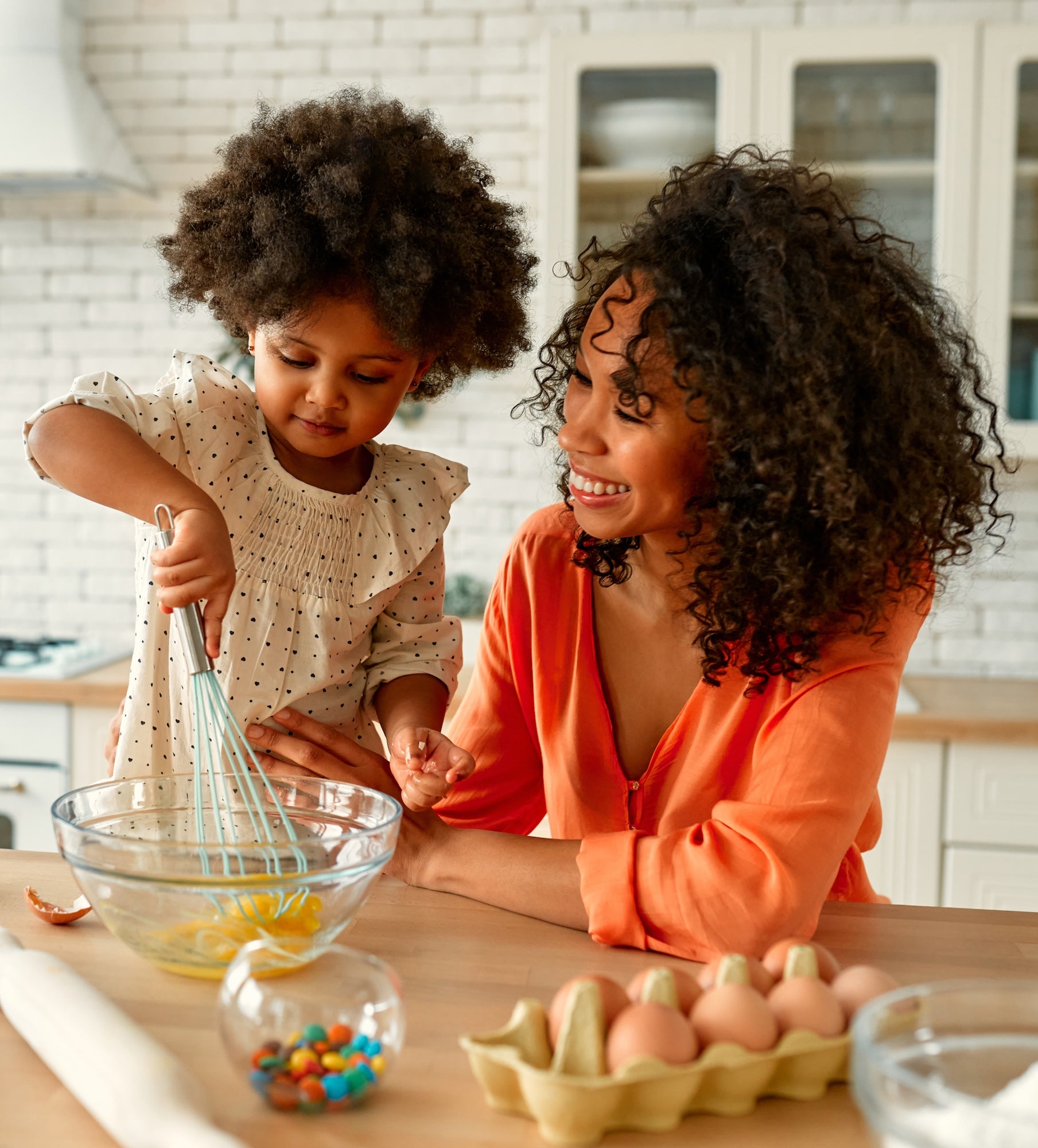 A smiling woman watches as a young child whisks eggs in a glass bowl on a kitchen counter. A carton of eggs, an egg shell, and a small bowl of colorful candies are nearby.