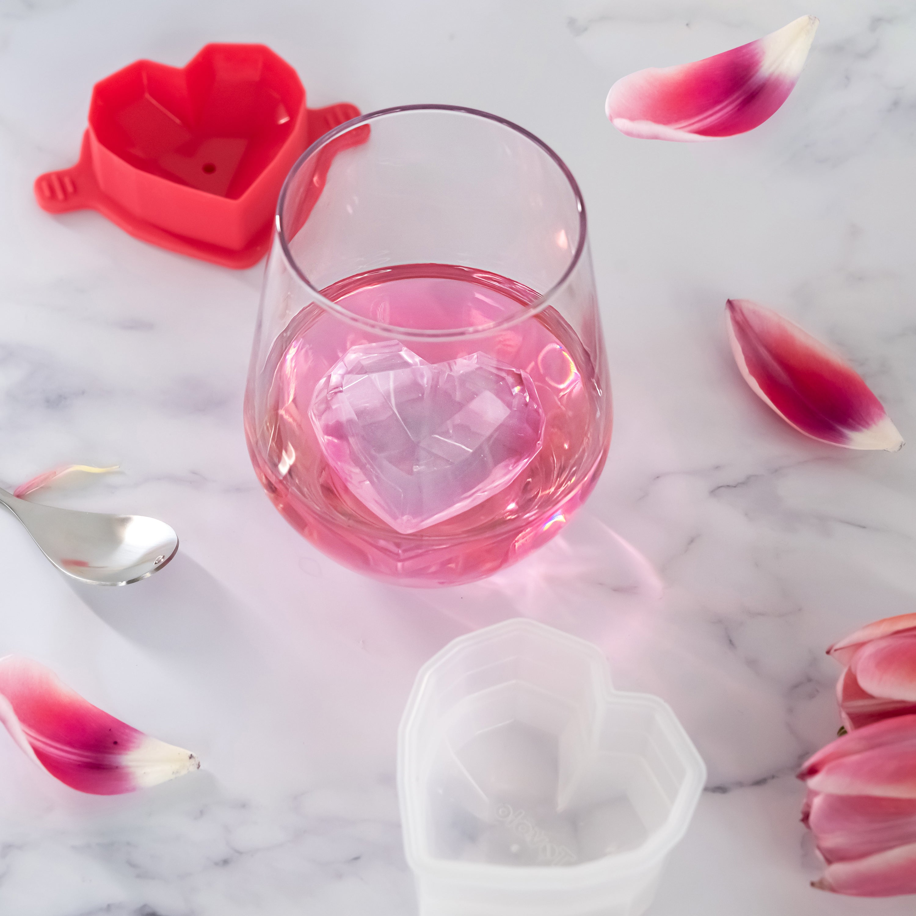 A glass of pink drink with a heart-shaped ice cube, surrounded by pink flower petals, a spoon, and heart-shaped silicone molds on a marble surface.