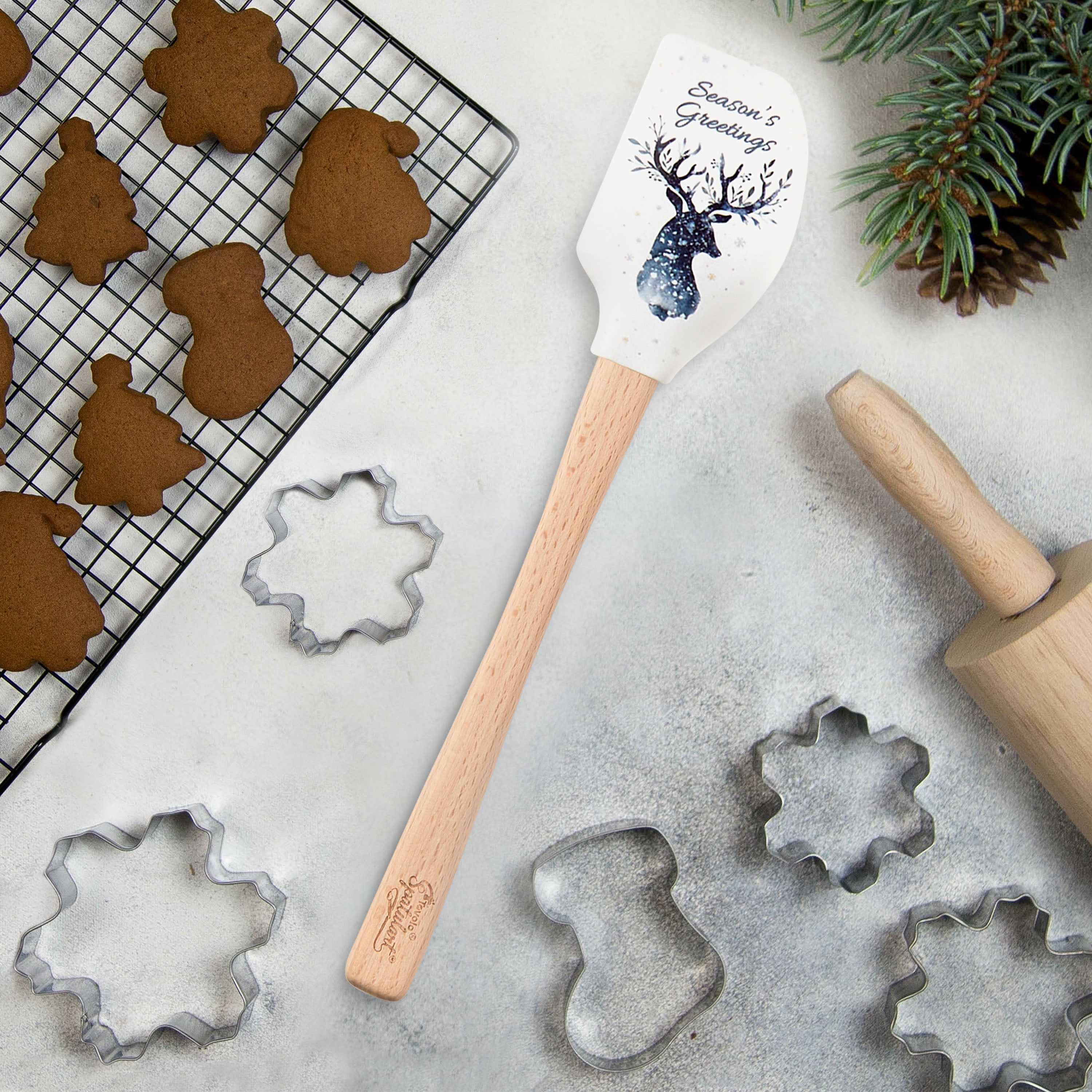A festive scene with iced gingerbread cookies shaped like a tree, deer, stars, and heart on parchment paper. A spatula with a tree pattern lies in the center, surrounded by cinnamon sticks and pine branches.