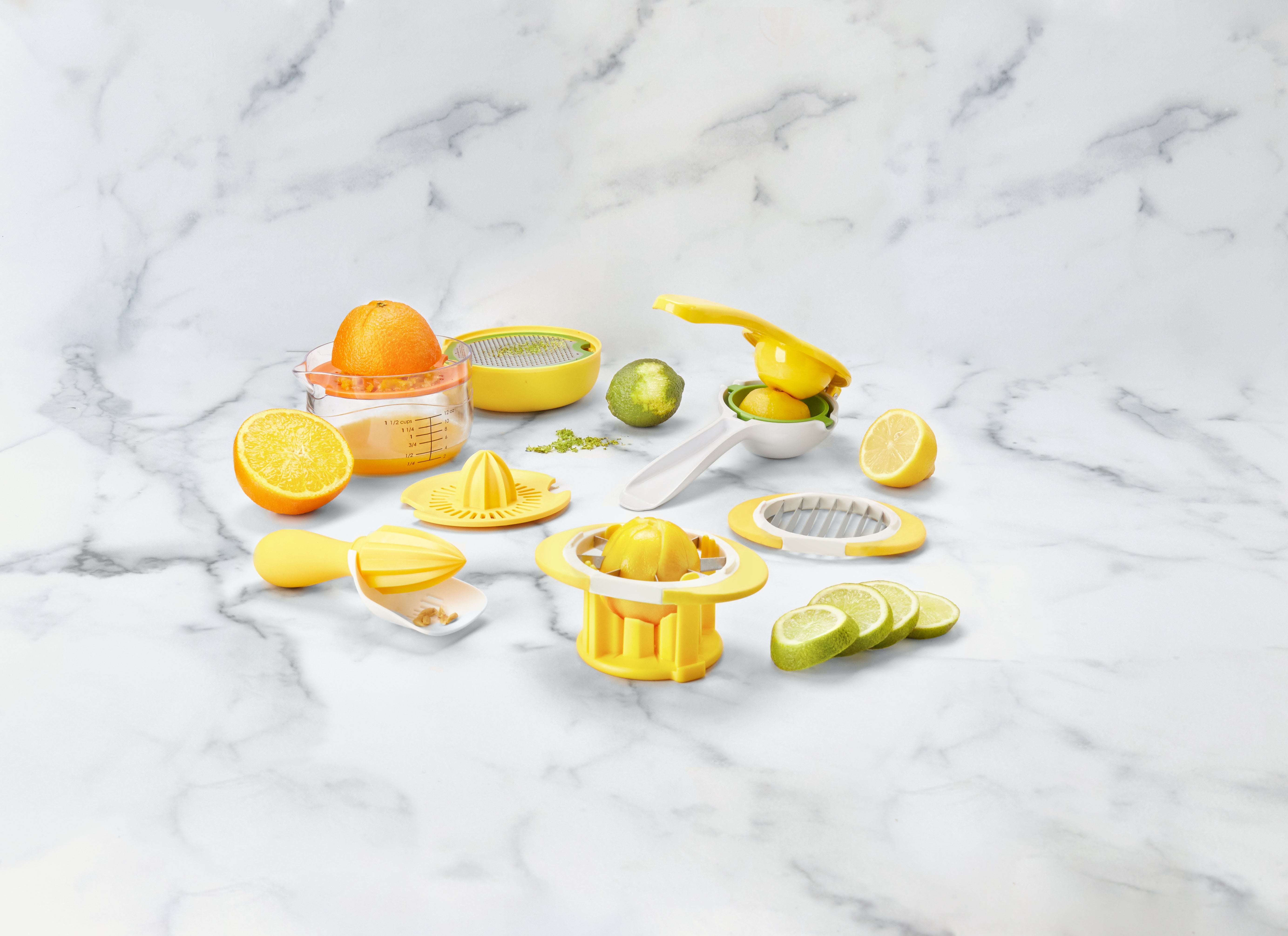 A variety of yellow citrus juicers and citrus fruits, including lemons and limes, are arranged on a white marble surface. Some fruits are sliced, and one orange is juiced in a measuring cup.