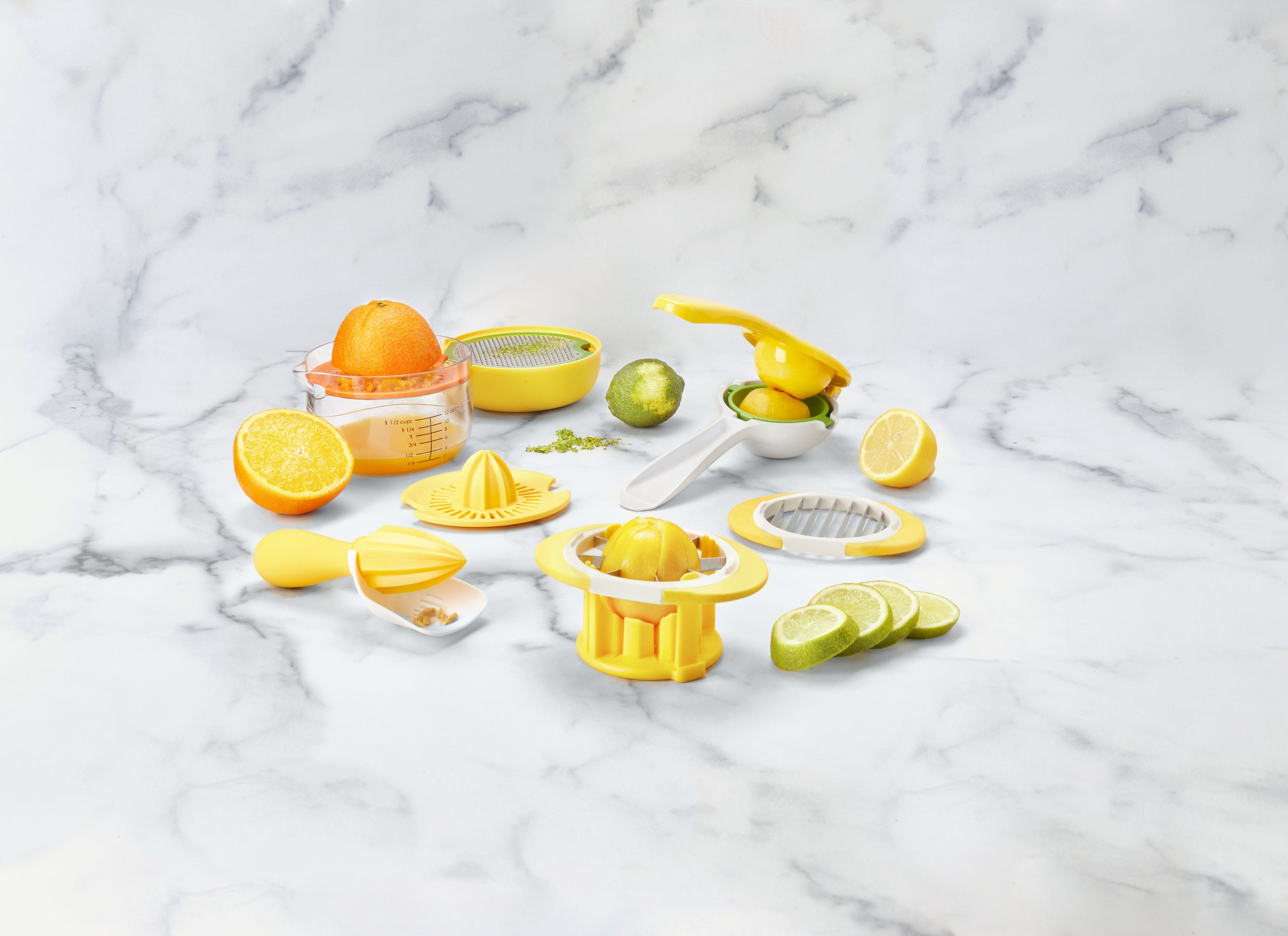 A variety of yellow citrus juicers and citrus fruits, including lemons and limes, are arranged on a white marble surface. Some fruits are sliced, and one orange is juiced in a measuring cup.