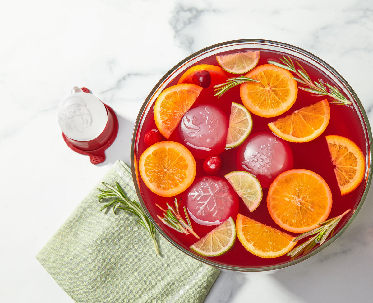 A bowl of red punch garnished with orange slices, lime wedges, cranberries, rosemary sprigs, and decorative ice cubes, next to a folded green napkin and a Santa-shaped container.