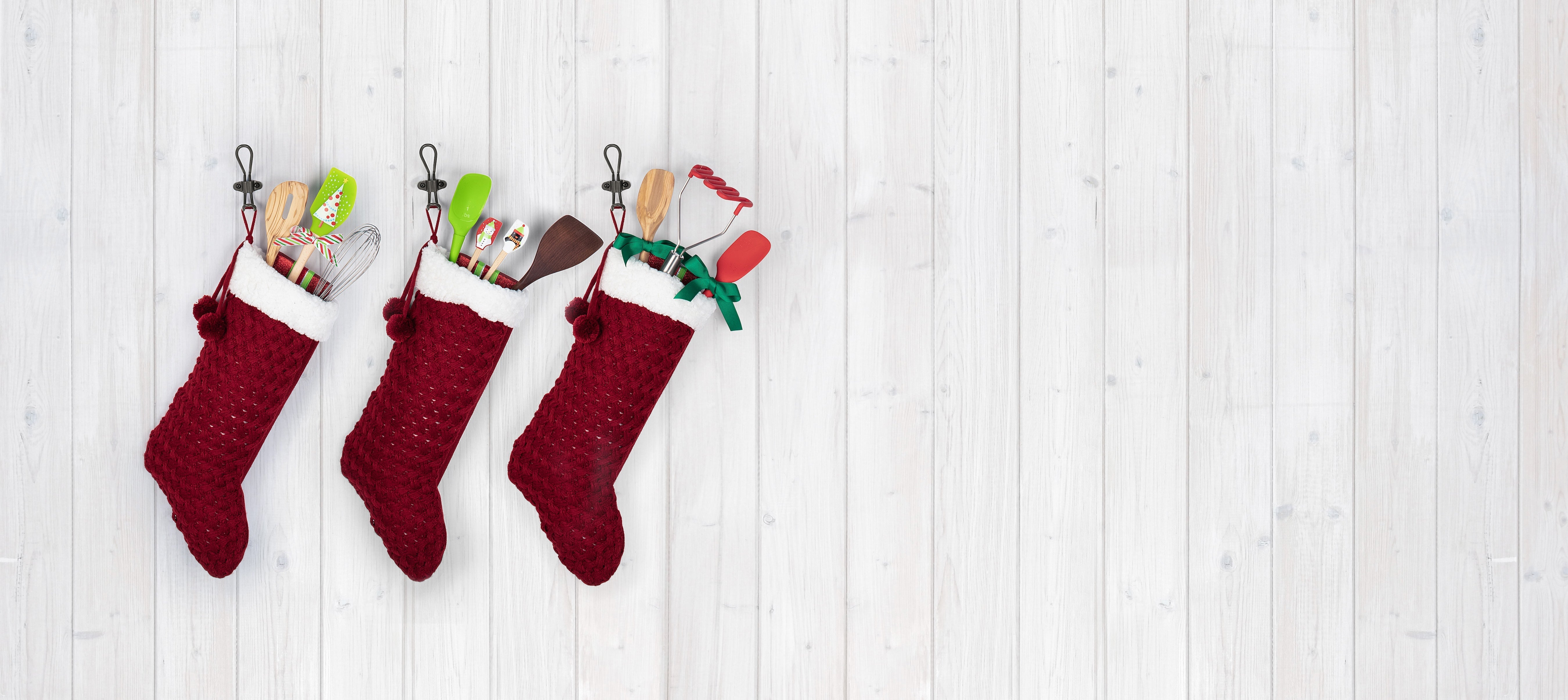 A wooden spatula with a stag design and Seasons Greetings text lies among metal cookie cutters, gingerbread cookies on a cooling rack, a rolling pin, and pine branches on a light surface.