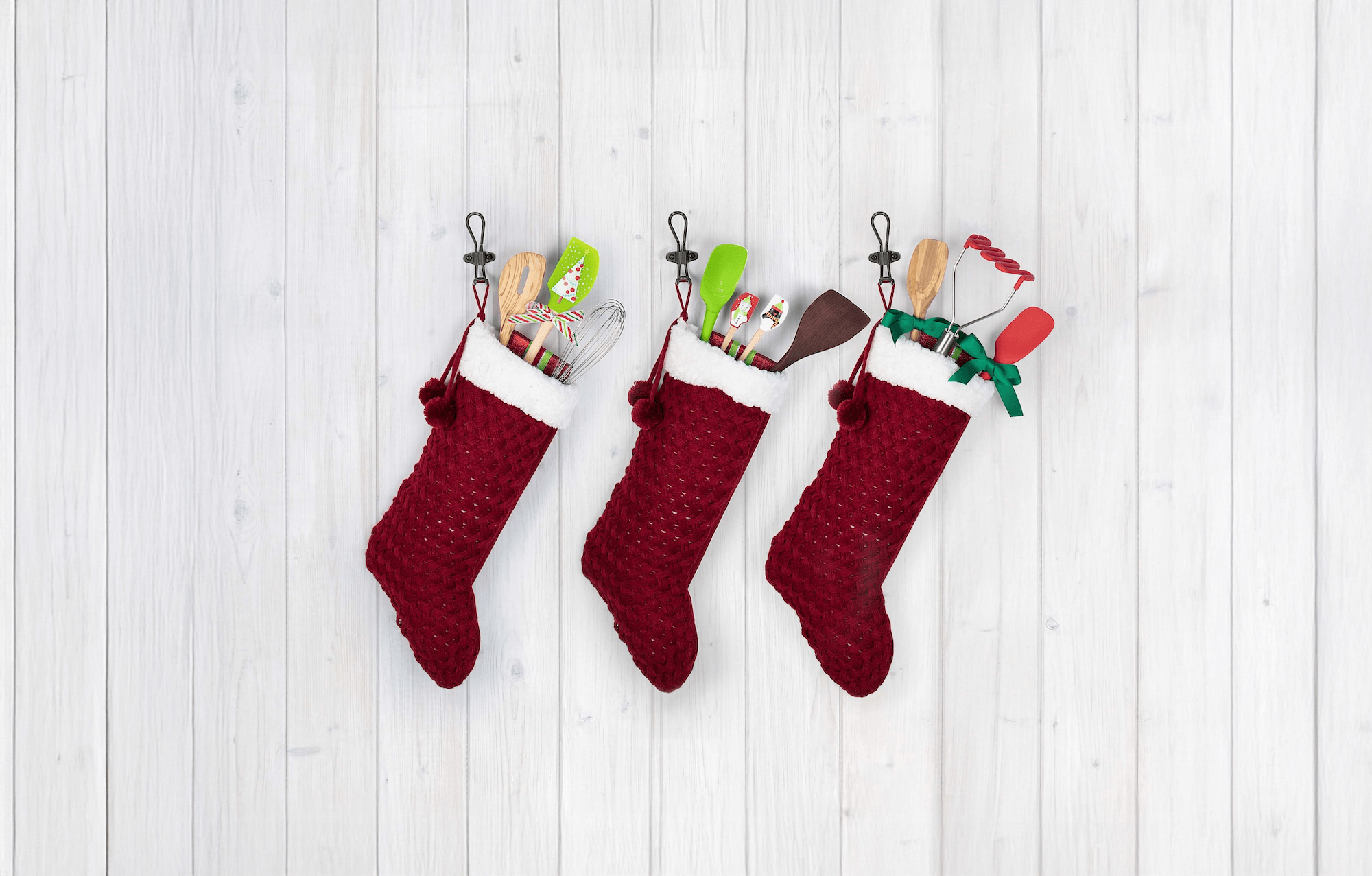 A wooden spatula with a stag design and Seasons Greetings text lies among metal cookie cutters, gingerbread cookies on a cooling rack, a rolling pin, and pine branches on a light surface.