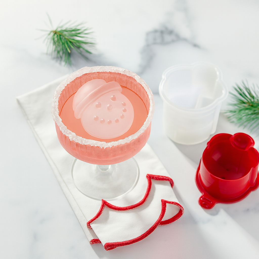 A pink cocktail in a sugar-rimmed glass with a snowman-shaped ice cube, napkin, ice mold pieces, and pine branches on a white marble surface.