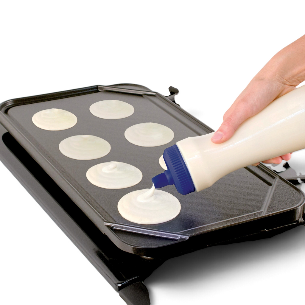 A hand uses the Pancake Pen 2.0 to squeeze pancake batter onto a griddle, creating several round pancakes. Burner controls are visible on the stove in front of the griddle.