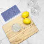 On a white marble surface: two lemons on a wooden cutting board, two empty glasses, and a blue Mini Ice Mold filled with mini ice cubes—ideal for instantly chilling drinks.