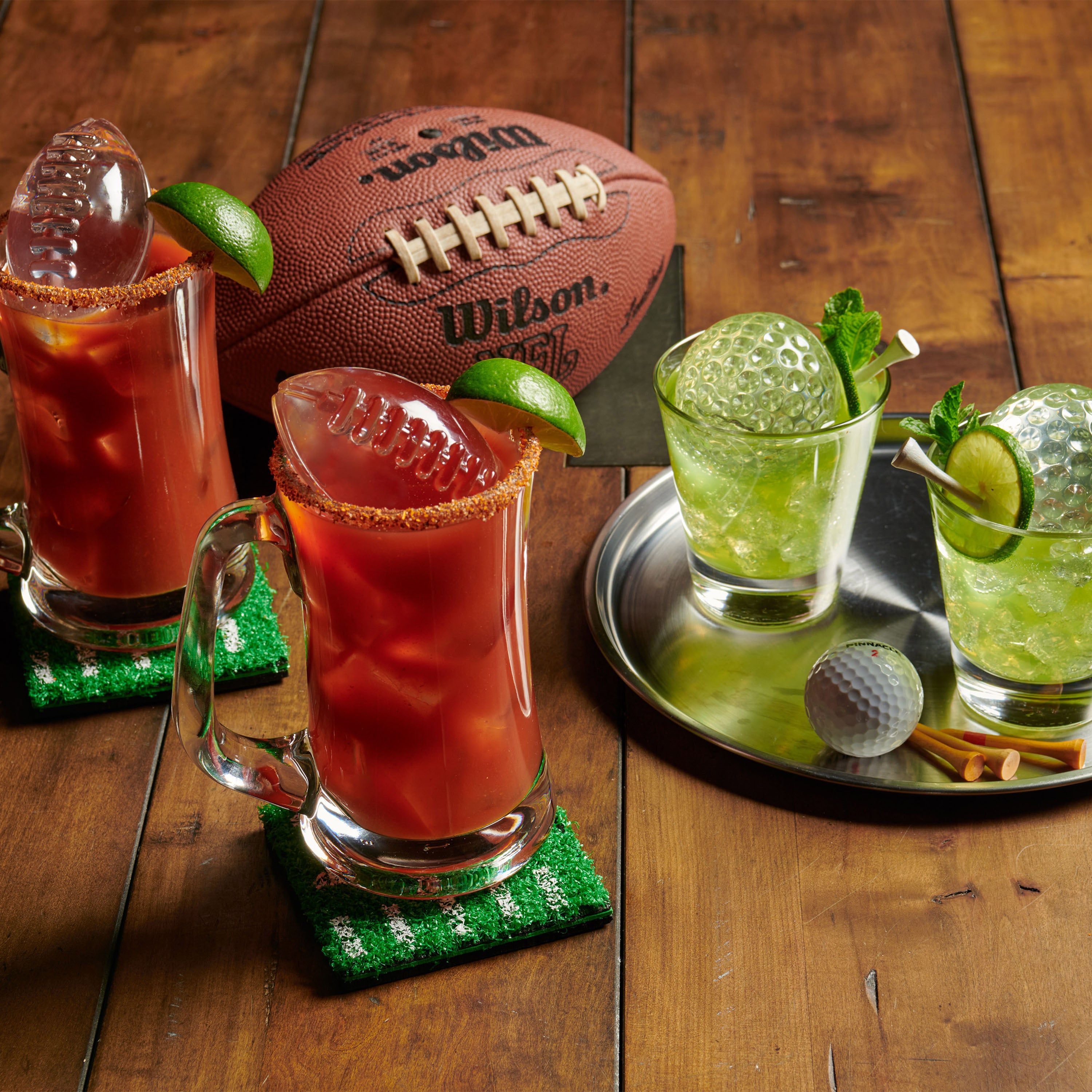 Two Bloody Marys and two mojitos with tovolo Golf and Football Sports Ice Molds (Set of 4) chill on a wooden table, with matching sports-themed coasters, tray, and football and golf ball decor.