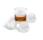 A glass of whiskey with a large, golf ball-shaped ice from the tovolo Golf and Tennis Ice Molds (Set of 4) sits beside three more molds on a white background.