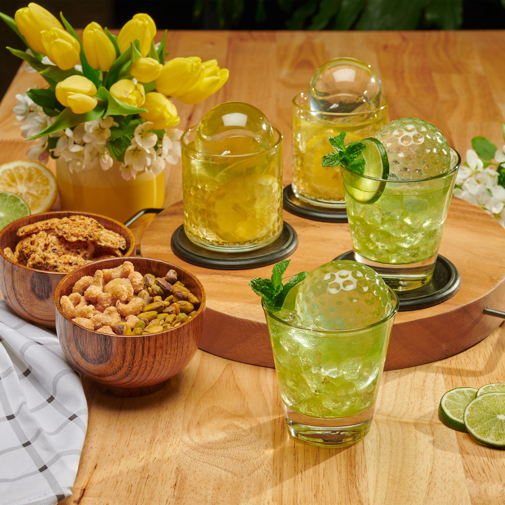 Two glasses of green cocktails with mint and lime sit on a wooden table, perfectly chilled with tovolo’s Golf and Tennis Sports Ice Molds (Set of 4), alongside snacks, sliced lime, a checkered napkin, and a vase of yellow tulips.