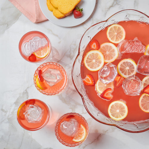 On a marble surface, a bowl and glasses of pink punch with ice made from the tovolo Hearts and Diamonds Ice Mold (Set of 4), lemon slices, and strawberries are displayed next to a plate holding yellow cake slices and a whole strawberry.