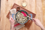 Using the tovolo Flex-Core Silicone and Stainless Steel Spoonula, someone stirs creamy pasta with red pepper flakes in a pot on a wooden board, next to a bowl of shredded cheese and a small cup of pepper flakes.