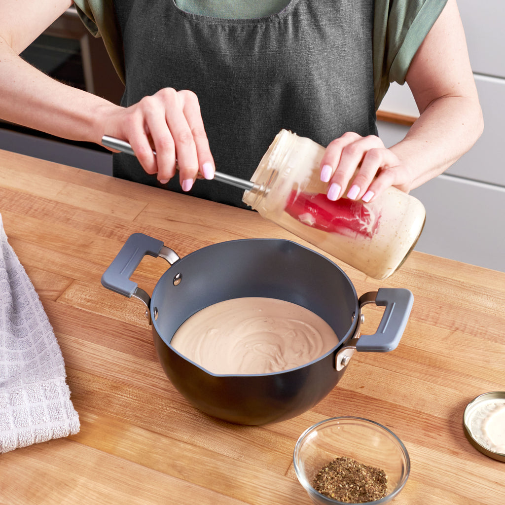 Wearing an apron, a person uses the tovolo Flex-Core Silicone and Stainless Steel Jar Scraper to pour creamy sauce from a glass jar into a black pot with a stainless steel handle on a wooden countertop beside spices and a towel.