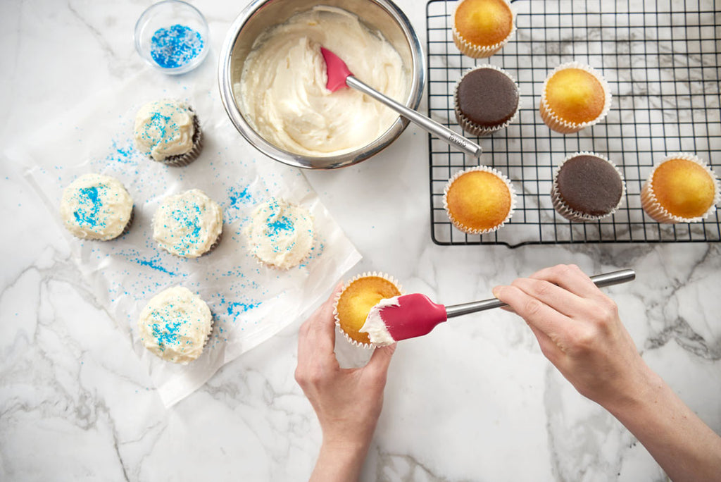 A person uses the tovolo Flex-Core Mini Spatula + Spoonula with Stainless Steel Handle to frost a cupcake. Nearby are frosted cupcakes with blue sprinkles, a bowl of frosting, blue sprinkles, and plain cupcakes on a cooling rack.