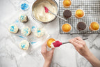 A person uses the tovolo Flex-Core Mini Spatula + Spoonula with Stainless Steel Handle to frost a cupcake. Nearby are frosted cupcakes with blue sprinkles, a bowl of frosting, blue sprinkles, and plain cupcakes on a cooling rack.