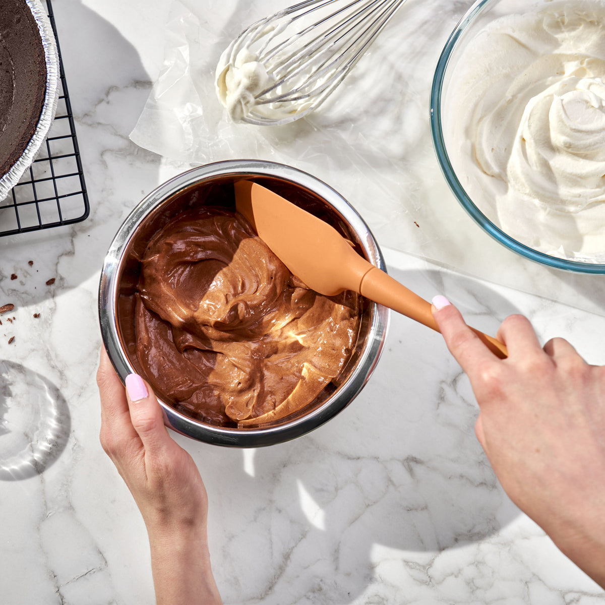 Using a tovolo Flex-Core Silicone Spatula, someone stirs creamy chocolate filling in a metal bowl on a marble counter. Nearby sit whipped cream, a whisk, and a pie crust cooling on a rack.