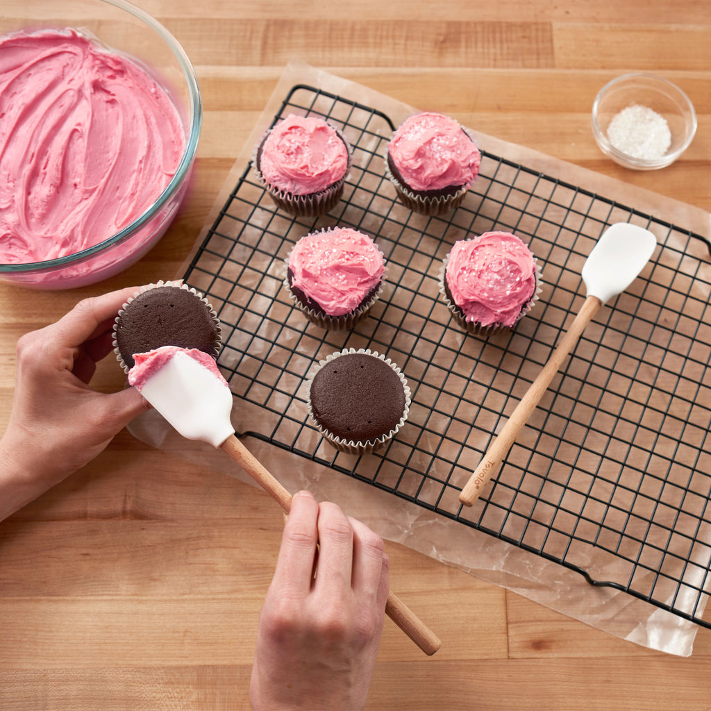 Someone uses the tovolo Flex-Core Mini Spatula + Spoonula with Wood Handle to spread pink frosting on a chocolate cupcake. More pink-frosted cupcakes with sprinkles sit on a rack beside a bowl of frosting and a dish of white sprinkles.
