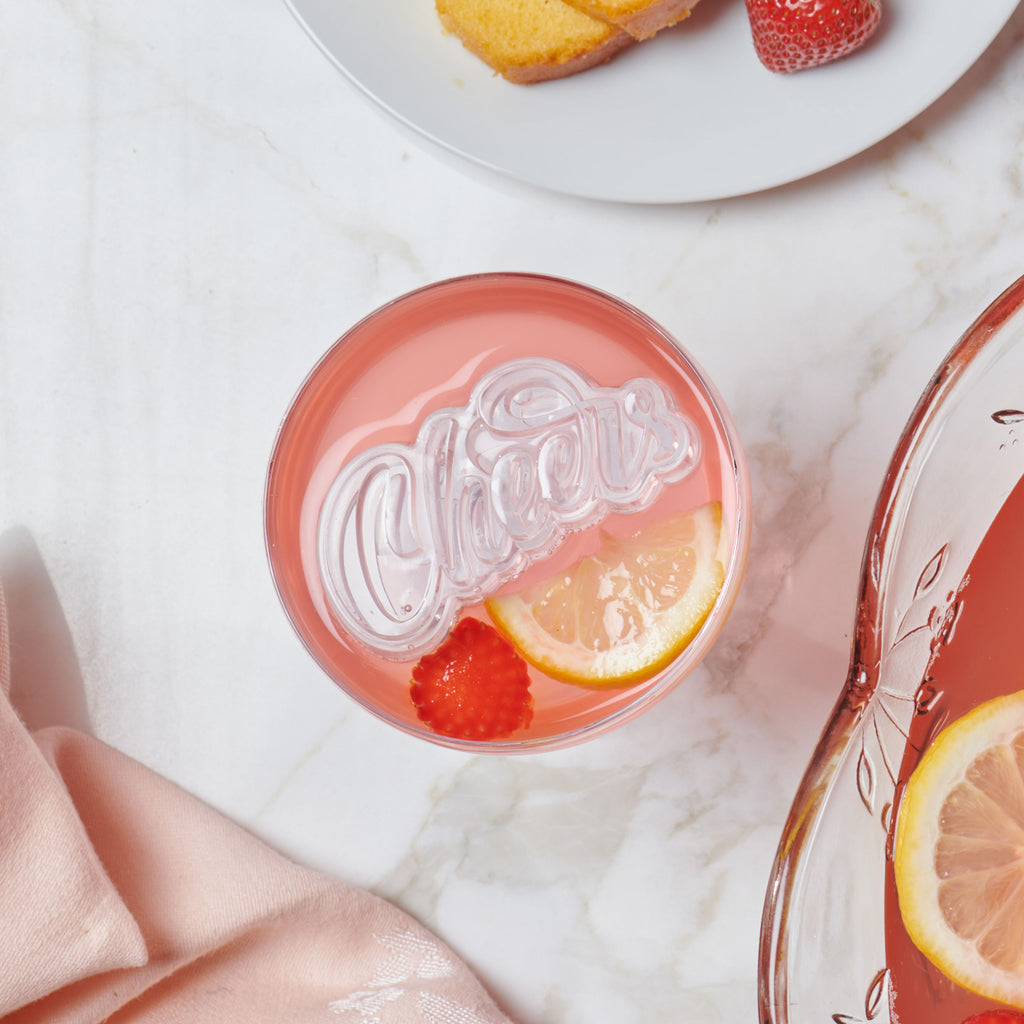 A pink drink with lemon and raspberry slices in a glass topped with a heart- or diamond-shaped ice cube from the tovolo Hearts and Diamonds Ice Mold set. Nearby are a pink napkin, punch bowl, and white plate with cake and a strawberry.