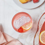 A pink drink with lemon and raspberry slices in a glass topped with a heart- or diamond-shaped ice cube from the tovolo Hearts and Diamonds Ice Mold set. Nearby are a pink napkin, punch bowl, and white plate with cake and a strawberry.