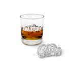 A glass of brown liquor with a large “Cheers” ice cube made using the tovolo Cheers Ice Mold (Set of 2) sits on a white surface, with another identical cube—also created with the tovolo silicone molds—resting beside it.