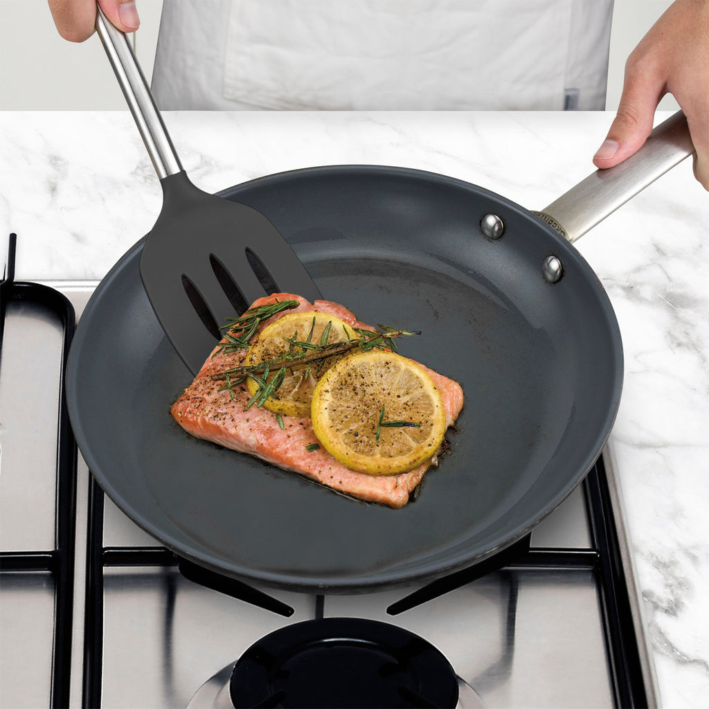 A person uses the tovolo Silicone and Stainless Steel Slotted Turner to cook salmon topped with lemon slices and herbs in a frying pan on a stovetop.