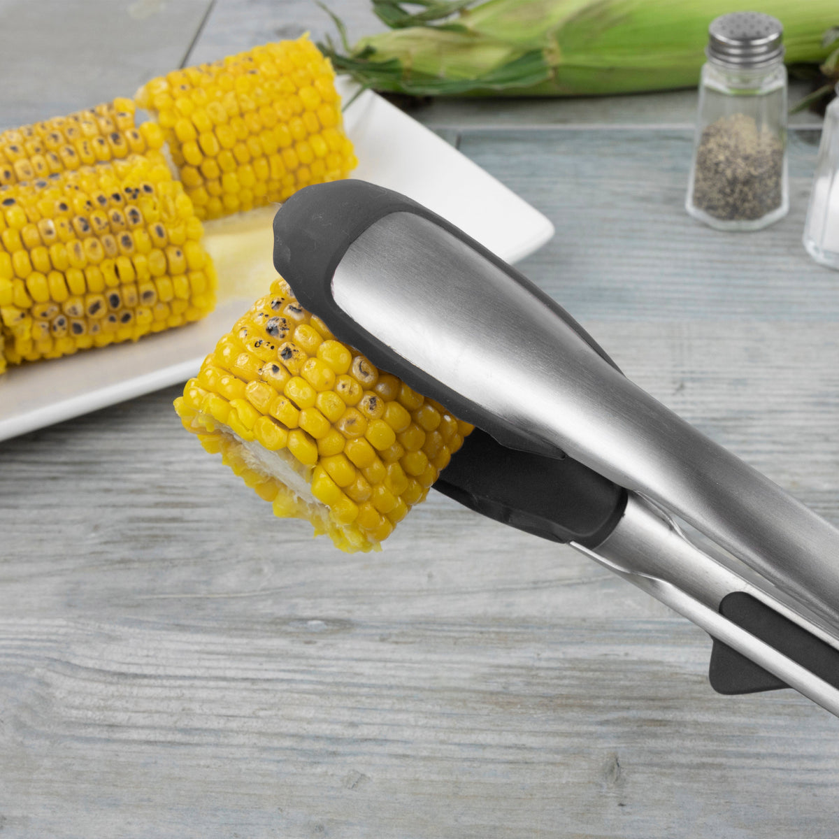 Tip Top Tongs holding cooked corn on the cob, with additional corn pieces on a white plate in the background, alongside salt and pepper shakers on a wooden surface.