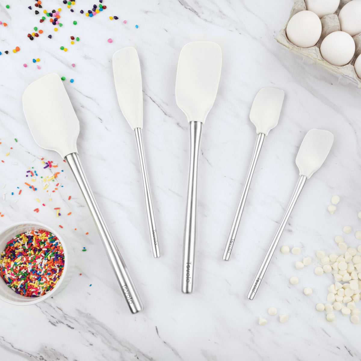 Five Flex-Core All-Silicone Spatula Set of 5 pieces in assorted sizes are displayed on a marble countertop with colorful sprinkles, white chocolate chips, and a carton of eggs.