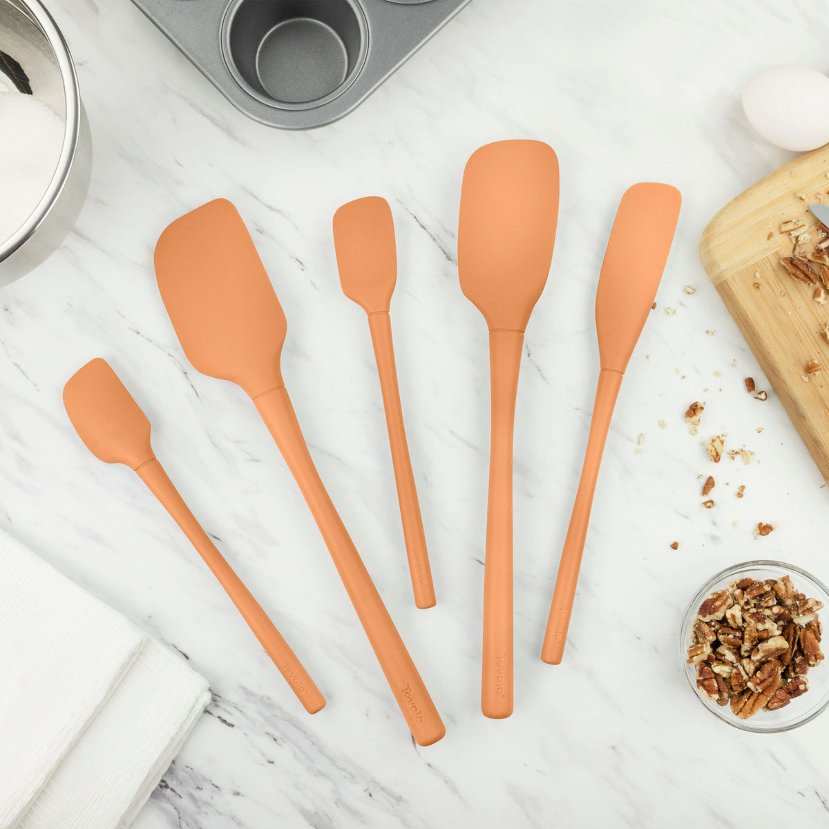 Five Flex-Core All-Silicone Spatula Set of 5 pieces in orange are arranged on a kitchen counter with a muffin tin, chopped pecans, a bowl of sugar, and a wooden cutting board.