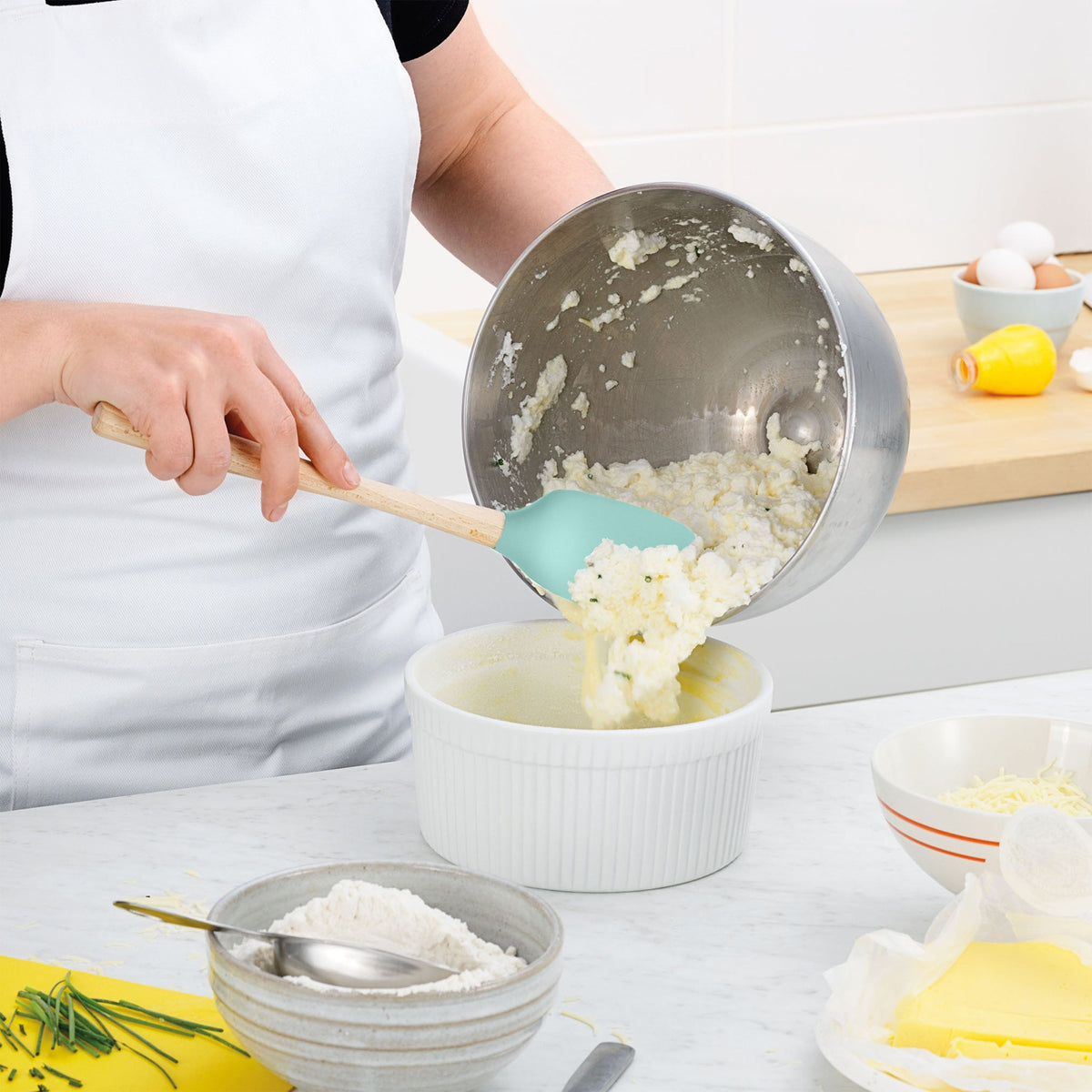 Wearing a white apron, someone uses a tovolo Flex-Core Silicone Spoonula to move a mixture from a metal bowl into a white baking dish, with flour, cheese, butter, and eggs displayed on the counter.