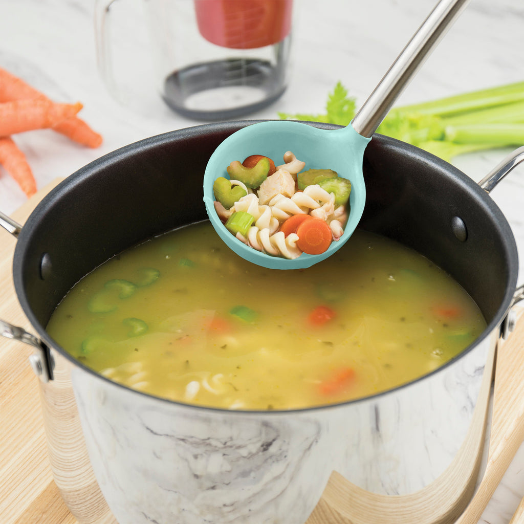 A pot of chicken noodle soup simmers on the stove. The tovolo Silicone & Stainless Steel Ladle serves up pasta, carrots, celery, and chicken, while fresh vegetables rest on the counter near the pot with its stainless steel handle.