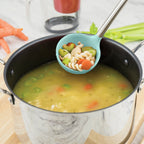 A pot of chicken noodle soup simmers on the stove. The tovolo Silicone & Stainless Steel Ladle serves up pasta, carrots, celery, and chicken, while fresh vegetables rest on the counter near the pot with its stainless steel handle.