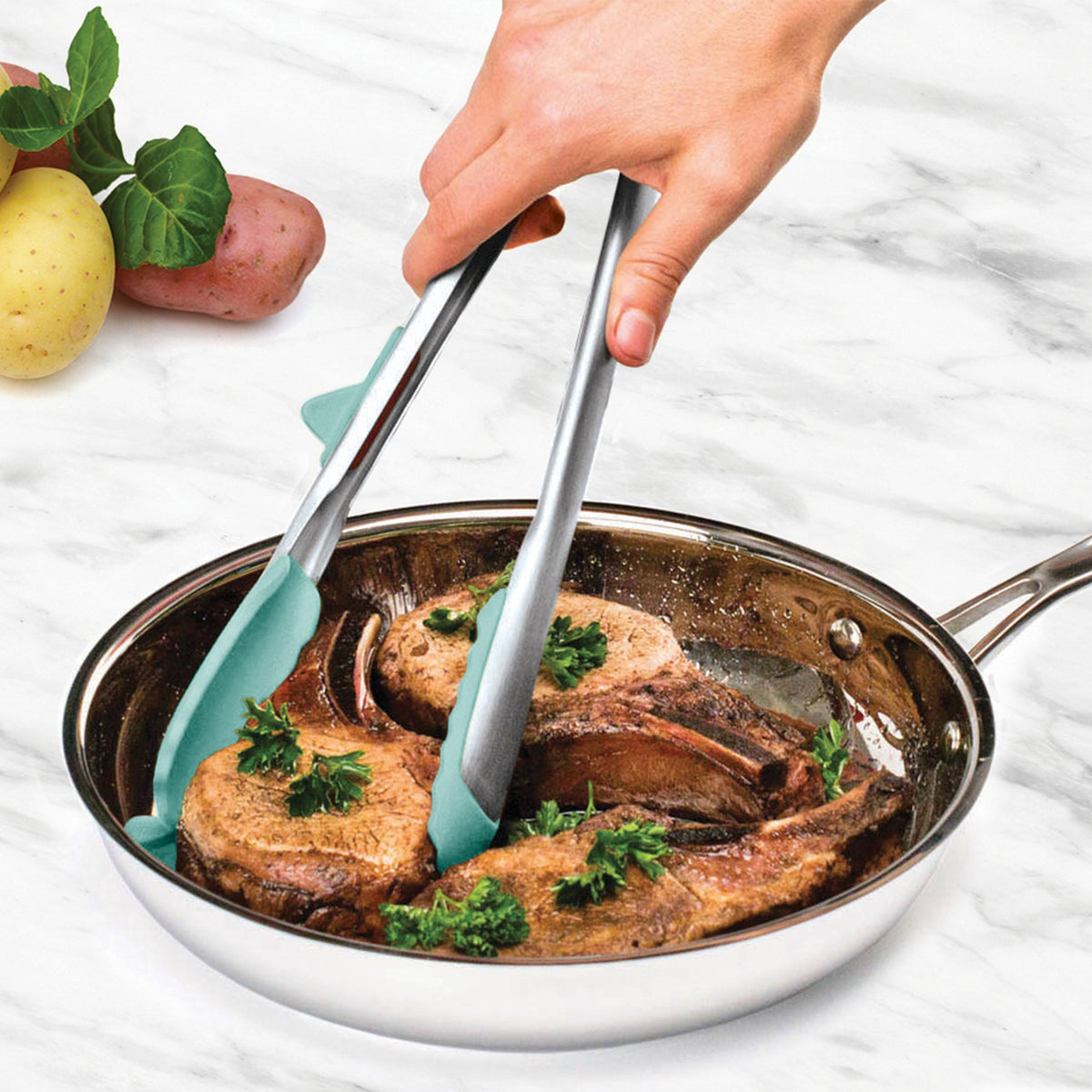 A hand uses Tip Top Tongs with a stainless steel handle to flip cooked pork chops garnished with parsley in a stainless steel skillet on a white marble countertop, with potatoes visible in the background.