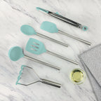 The tovolo Silicone and Stainless Steel Utensil Set of 6 features heat-resistant, non-scratch teal silicone heads and stainless steel handles, displayed on white marble beside a glass bowl of oil and a gray textured cloth.