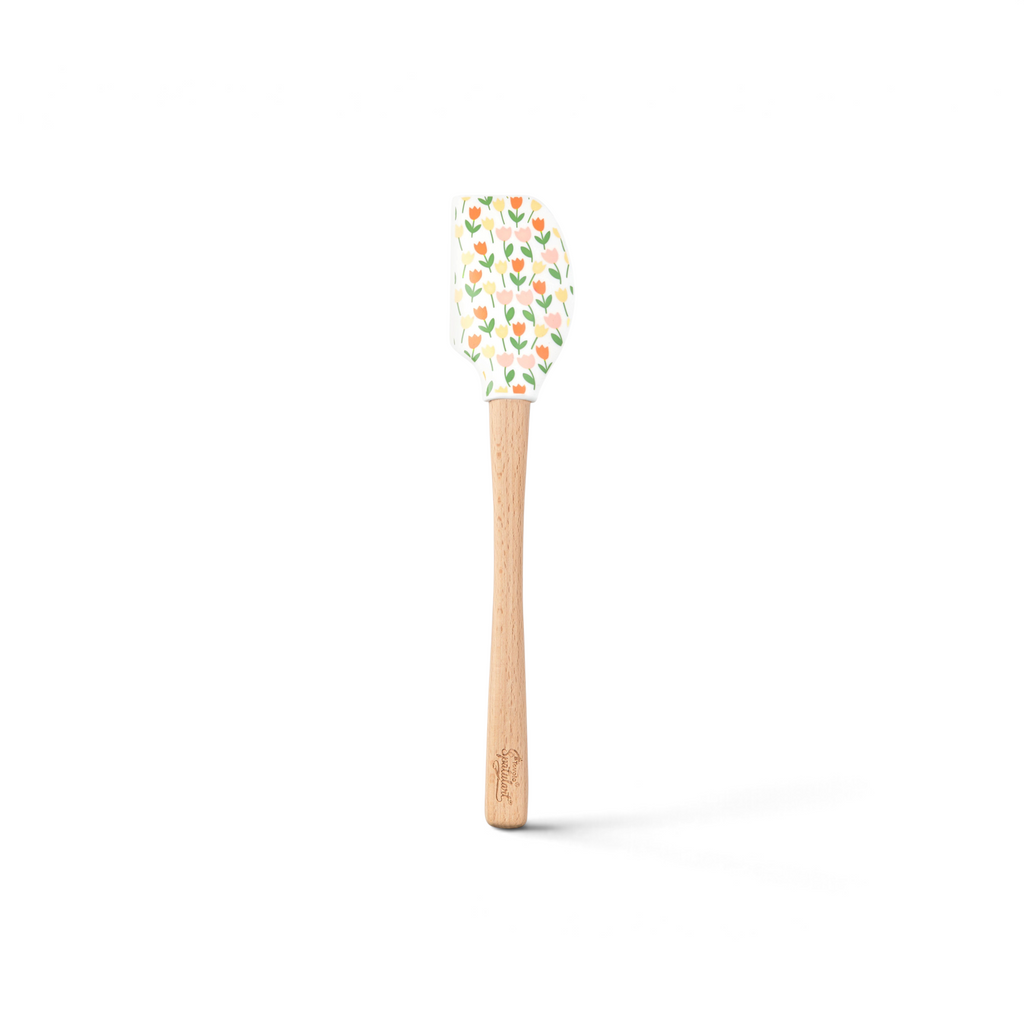 The Spring Spatulart is a wood-handled silicone spatula featuring a carrot patch design, with orange and yellow tulips and green leaves on the spatula head.