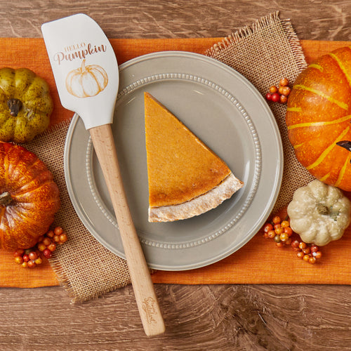 A slice of pumpkin pie on a beige plate sits on an orange placemat with small pumpkins and berries. Nearby rests the Autumn Spatulart, a wood-handled spatula featuring “Hello Pumpkin” and a pumpkin illustration.