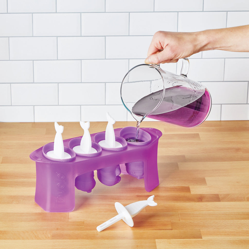 A hand pours purple liquid into Pop Molds with white whale-shaped sticks on a wooden countertop, making homemade frozen treats in front of a white tile wall.